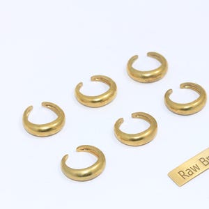 May include: Six gold-colored brass ear cuffs are arranged on a white surface. The ear cuffs have a curved, open design. A small rectangular tag with the text "Raw Brass" is also present.