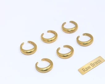 May include: Six gold-coloured brass ear cuffs are arranged on a white surface. The ear cuffs have a curved, open design. A small rectangular tag with the text "Raw Brass" is also present.