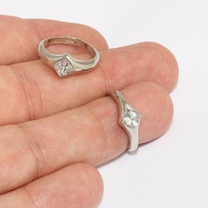 May include: Two silver-toned rings with a square, clear gemstone. One ring is on a finger, the other is adjustable. The rings have a simple, elegant design, perfect for everyday wear. The rings are set against a white background.