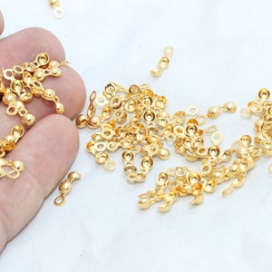 Puede incluir: Primer plano de hallazgos de joyería dorados. Estos pequeños componentes decorativos presentan una serie de cuentas y bucles conectados, ideales para la fabricación de joyas. Los hallazgos están dispersos sobre una superficie blanca.