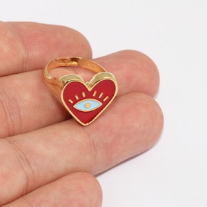 May include: Gold-toned adjustable ring featuring a red heart-shaped face with a blue eye design. The ring is held in a hand against a white background. The heart has a gold outline.