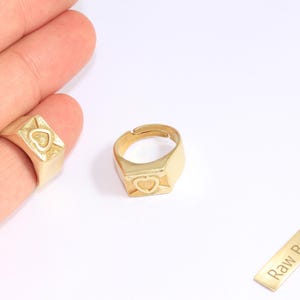 以下が含まれることがあります： ハートのデザインが施された、2つのゴールドカラーのシグネットリング。1つのリングは指にあり、もう1つは調整可能です。「Raw Brass」の文字が入った小さな真鍮の長方形も存在します。リングは白い背景にあります。