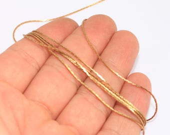 May include: Close-up of several strands of gold-colored chain. The chains are thin and delicate, with a slightly textured surface. The chains are held in a hand against a white background. The chains appear to be new.