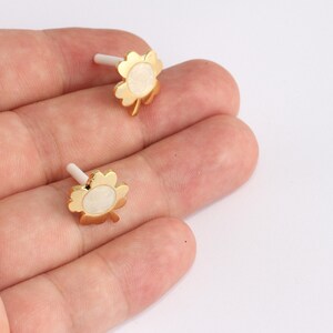 May include: Two gold-coloured clover-shaped earrings with white centres and white posts. The earrings are displayed on a light background. The earrings are small and delicate, with a shiny finish.