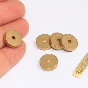 Könnte beinhalten: Eine Sammlung von Rohmessingkomponenten, darunter mehrere kleine, runde, flache Scheiben mit einem zentralen Loch und ein rechteckiges Etikett mit dem Text "Raw Brass". Die Komponenten haben eine warme Goldfarbe.