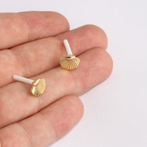 Puede incluir: Dos pendientes dorados con forma de concha marina y postes blancos. Los pendientes tienen un diseño detallado en forma de abanico. Los pendientes son pequeños y delicados, adecuados para el uso diario o ocasiones especiales.