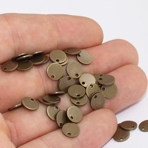 以下が含まれることがあります： 小さな丸いアンティーク真鍮色の金属チャームのコレクション。上部に1つの穴があります。チャームは明るい背景に散らばっており、ジュエリー作りやクラフトプロジェクトでの使用を示唆しています。