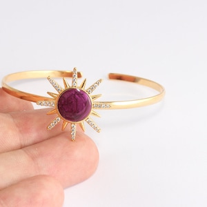May include: Gold-tone cuff bracelet featuring a sunburst design. The sunburst has a deep purple center and is surrounded by sparkling clear crystals. The bracelet is open-ended for easy wear and adds a touch of celestial elegance.