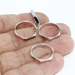 Puede incluir: Un conjunto de anillos geométricos de color plateado. Los anillos tienen forma hexagonal con lados planos y un acabado pulido. La imagen muestra los anillos en varias orientaciones, con un anillo parcialmente visible.