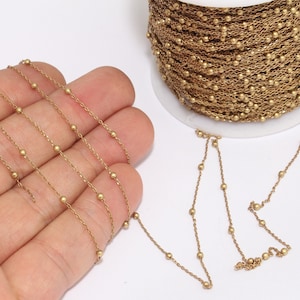 May include: A spool of gold-coloured beaded chain. The chain is made of small, round beads connected by delicate links. A length of the chain is unspooled, with some of it held in a hand. The chain is on a white background.