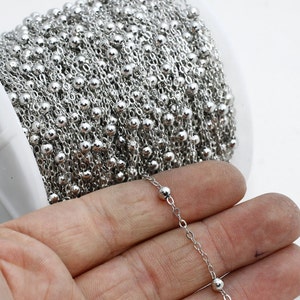 Könnte beinhalten: Eine Rolle glänzender silberner Kugelkette. Die Kette besteht aus kleinen, runden Perlen, die durch zarte Glieder verbunden sind. Die Kette ist eng auf einer weißen Spule aufgewickelt, wobei ein Teil der Kette herausragt.