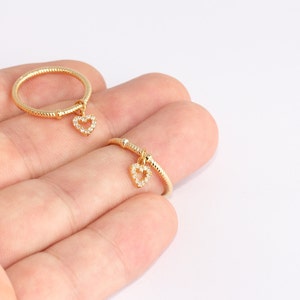 May include: Two gold-colored rings with a heart-shaped charm. The heart charms are encrusted with small, clear stones. The rings have a textured band design. The rings are displayed on a light background.