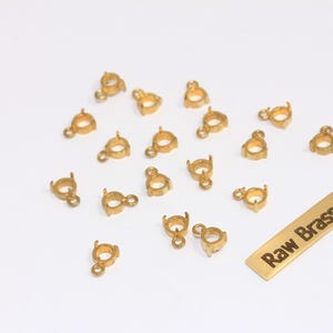 May include: A collection of small, gold-colored jewelry findings, including bezel settings with loops. A rectangular tag with the text "Raw Brass" is also present. The findings are scattered on a white surface.