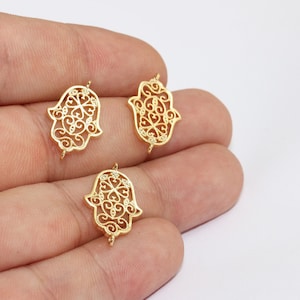 Puede incluir: Tres colgantes dorados con forma de mano de Hamsa, con intrincados diseños de filigrana. Cada colgante presenta un detallado patrón calado y pequeños bucles para sujetar a la joyería. Los colgantes se muestran sobre un fondo neutro.
