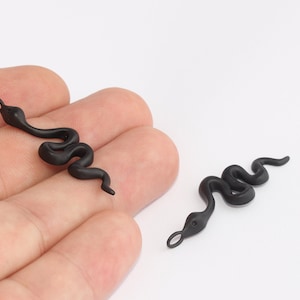 May include: Two matte black snake charms. Each charm is a stylized representation of a snake, with a curved body and a small loop at the head. The charms are designed for jewellery making or crafting.