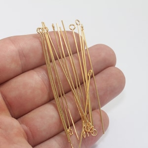 Puede incluir: Una colección de alfileres dorados con lazos y alfileres rectos. Los alfileres son finos y parecen ser de metal. Los alfileres se sostienen en una mano sobre un fondo blanco. Estos alfileres se utilizan para la fabricación de joyas y manualidades.