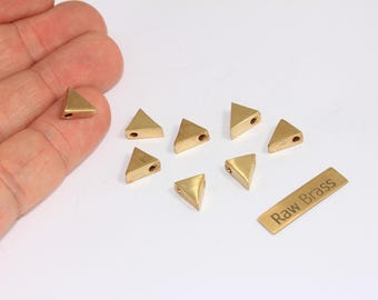 May include: A collection of small, triangular brass beads with a hole drilled through each. The beads are a warm gold color and are arranged on a white surface. A small brass tag with the text "Raw Brass" is also present.