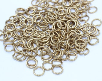 Puede incluir: Un montón de pequeños anillos abiertos de color latón cálido. Estos anillos metálicos se utilizan para la fabricación de joyas y otras manualidades. Los anillos están dispersos sobre una superficie blanca.