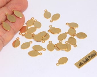 May include: A collection of small, gold-plated oval charms with a loop at the top for jewelry making. The charms are scattered on a white surface, with a small rectangular tag that reads "24k Gold Plated".
