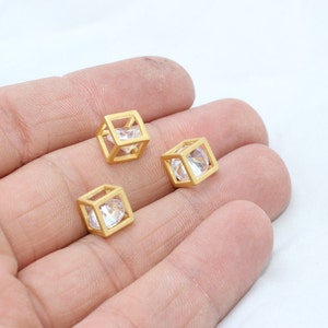 以下が含まれることがあります： 透明なファセットストーンを中央に配置した、金色の立方体型のビーズが3つ。白い背景に、手でビーズが持たれています。立方体は、開いた幾何学的なデザインです。