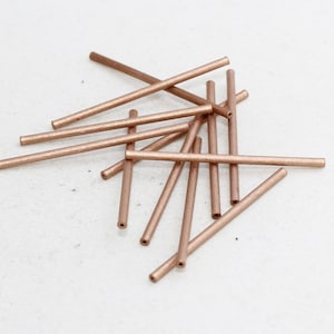 Puede incluir: Un conjunto de tubos metálicos de color cobre dispersos sobre una superficie blanca. Los tubos son cilíndricos, con un centro hueco y una superficie ligeramente texturizada. La iluminación resalta el brillo metálico.