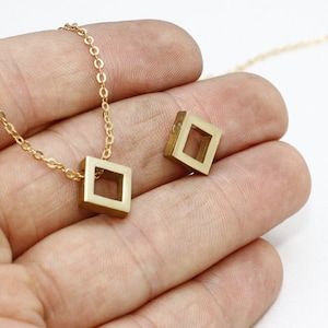 May include: Gold-colored square pendant necklace and a matching charm. The pendant is a hollow square, suspended from a delicate chain. The charm is a similar square shape. The jewelry is displayed against a neutral background.