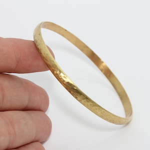 May include: A hammered gold-tone bangle bracelet. The bracelet has a textured surface and a circular shape. The gold color is bright and shiny. The bangle is held by a hand against a white background. Jewelry.