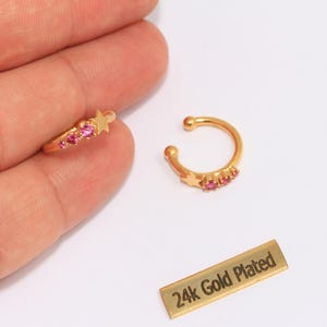 May include: Two gold-plated ear cuffs with pink gemstones and star accents. One cuff is on a finger, the other is separate. A small gold-plated rectangle reads "24k Gold Plated".