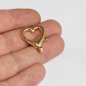 Könnte beinhalten: Ein goldfarbener herzförmiger Verschluss. Der Verschluss besteht aus Metall und hat einen federbelasteten Mechanismus. Die Herzform ist offen, mit einem geschwungenen Design. Der Verschluss ist für Schmuck oder andere Bastelprojekte konzipiert.