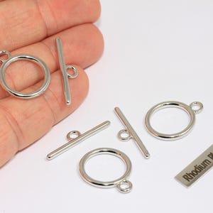 May include: Silver-colored toggle clasps, including round rings and bar components, are displayed. The clasps are arranged on a white surface, with one piece labeled "Rhodium Plated". These jewelry findings are for crafting.