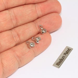 May include: Three silver-coloured, bell-shaped jewellery findings with small loops, displayed on a hand. A small rectangular tag with the text "Rhodium Plated" is also present. These findings are likely used for jewellery making.