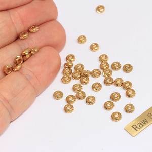 May include: Small, gold-coloured raw brass beads with a decorative pattern are scattered on a white surface, with some held in a hand. A small brass tag reads "Raw Brass".