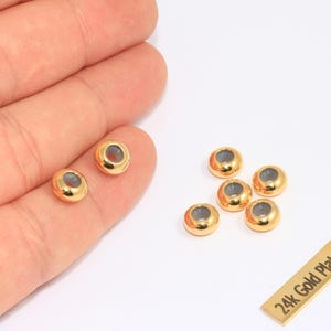 Könnte beinhalten: Vergoldete Perlen mit hohler Mitte, präsentiert auf einer weißen Oberfläche. Die Perlen haben eine glänzende, reflektierende Oberfläche. Ein kleines rechteckiges Etikett lautet "24k Gold Plated".