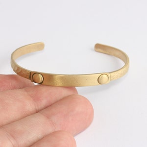 Może przedstawiać: Złota bransoletka na nadgarstek z młotkowaną fakturą na zewnętrznych krawędziach i gładkim, prostokątnym środkiem. Bransoletka posiada dwa okrągłe akcenty. Biżuteria.