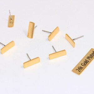 May include: A collection of gold-plated bar stud earrings. The earrings are rectangular with a post for piercing. One bar has the text "24k Gold Plated". The earrings are scattered on a white surface.