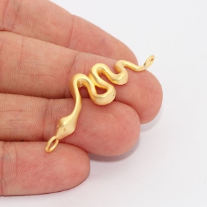Könnte beinhalten: Ein goldfarbener Schlangenanhänger mit geschwungenem Körper und einer kleinen Schlaufe an jedem Ende. Der Schlangenkopf hat ein kleines Augendetail. Ideal für die Herstellung von Halsketten, Armbändern oder Ohrringen.