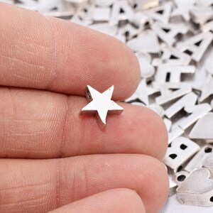 Könnte beinhalten: Eine kleine, silberfarbene sternförmige Perle wird zwischen den Fingern gehalten, mit einer Sammlung anderer Metallbuchstaben und Formperlen im Hintergrund. Die Perlen sind wahrscheinlich für die Schmuckherstellung oder Bastelprojekte bestimmt.
