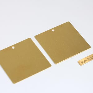 Könnte beinhalten: Zwei quadratische, goldfarbene Metallanhänger mit einem kleinen Loch oben und ein kleines rechteckiges Etikett mit der Aufschrift "Raw Brass". Die Anhänger befinden sich auf weißem Hintergrund.
