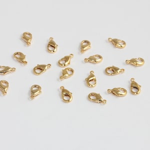 Puede incluir: Una colección de pequeños cierres de langosta dorados, dispersos sobre una superficie blanca. Estos cierres se utilizan en la fabricación de joyas para asegurar collares, pulseras y otros accesorios. Los cierres son de metal y tienen un acabado brillante.