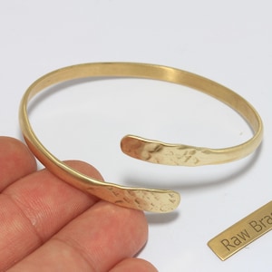 Op de afbeelding: Een goudkleurige open manchetarmband met gehamerde textuur aan de uiteinden. De armband is gemaakt van onbewerkt messing. Een klein rechthoekig label met de tekst "Raw Brass" is ook zichtbaar.