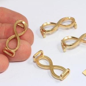 Può includere: Ciondoli a forma di simbolo dell'infinito color oro, possibilmente per la creazione di gioielli. I ciondoli hanno un anello su ciascuna estremità. È presente anche una piccola etichetta in ottone con la scritta "Raw Brass".