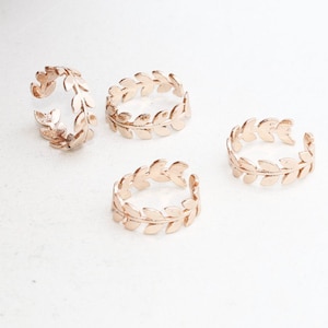 May include: Four rose gold adjustable rings with a leaf design are displayed on a white surface. The rings are open-ended, allowing for size adjustment. The leaf pattern wraps around the ring band.