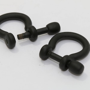 Könnte beinhalten: Zwei schwarze D-Ring-Schäkel mit Gewindestiften, präsentiert auf einer weißen Oberfläche. Ein Schäkel ist geöffnet und zeigt den Gewindestift, während der andere geschlossen ist. Die Schäkel bestehen aus mattem schwarzem Material.