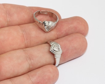 May include: Two silver-colored rings with heart-shaped designs. The rings have a ribbed band and a heart-shaped setting. One ring is on a finger, and the other is next to it. The rings are adjustable.
