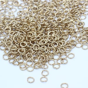 May include: A pile of small, gold-coloured jump rings scattered on a white surface. The rings are circular with a small opening, suggesting they are for jewellery making or crafts. The lighting highlights the metallic sheen.