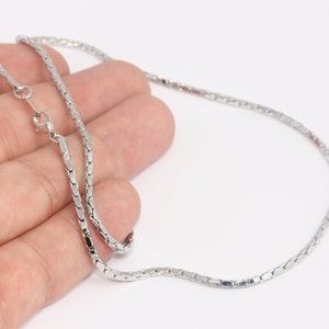 Può includere: Una collana color argento con una delicata catena a maglie rettangolari. La collana è mostrata su uno sfondo bianco e presenta una chiusura a fermaglio. La catena è tenuta da una mano.