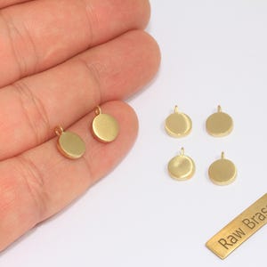 May include: A collection of small, gold-colored brass pendants in various shapes. Some are round, while others are oval. The pendants have a loop at the top for attaching to a chain or other jewelry component. A small brass rectangle with the text "Raw Brass" is also present.