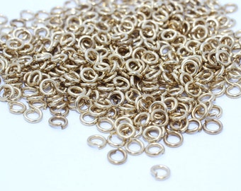 May include: A pile of small, gold-colored jump rings, scattered across a white surface. The rings are circular with a small opening, suggesting they are for jewelry making or crafts. The lighting highlights the metallic sheen.