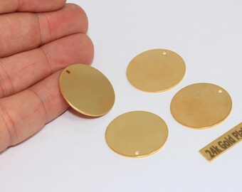 May include: Four circular, flat, gold-plated metal charms, each featuring a small hole near the top edge for attachment. A small rectangular tag is present with the text 24k Gold Plated printed in black on a gold background.