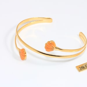 May include: Gold-plated cuff bracelet with two orange flower-shaped accents. The bracelet has a double-band design and is open-ended. A small rectangular tag reads "24k Gold Plated".